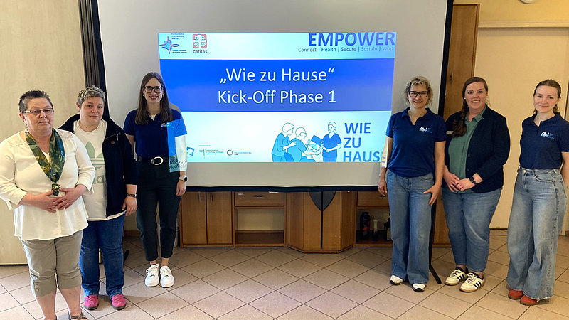 Links und rechts einer Beamer-Leinwand auf der der Projektname "Wie zu Hause " zu lesen ist, stehen jeweils drei Frauen und lachen in die Kamera.