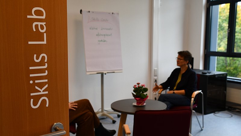 Blick durch die geöffnete Tür in das Skills Lab der KH Mainz. Im Hintergrund sind unscharf Personen in einer Beratungssituation zu sehen. 