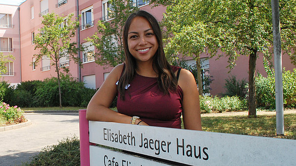 Janine Hänichen steht an einem Schild mit der Aufschrift Elisabeth Jäger Haus, einer Senioreneinrichtung der Stiftung kreuznacher diakonie. Das Haus ist im Hintergrund zu sehen.
