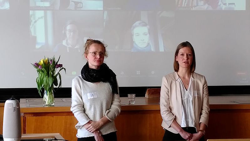 Zwei Mitarbeiterinnen aus dem Projekt FAMOUS stehen vor einer Leinwand, auf der andere Teilnehmende per zoom zugeschaltet sind. 