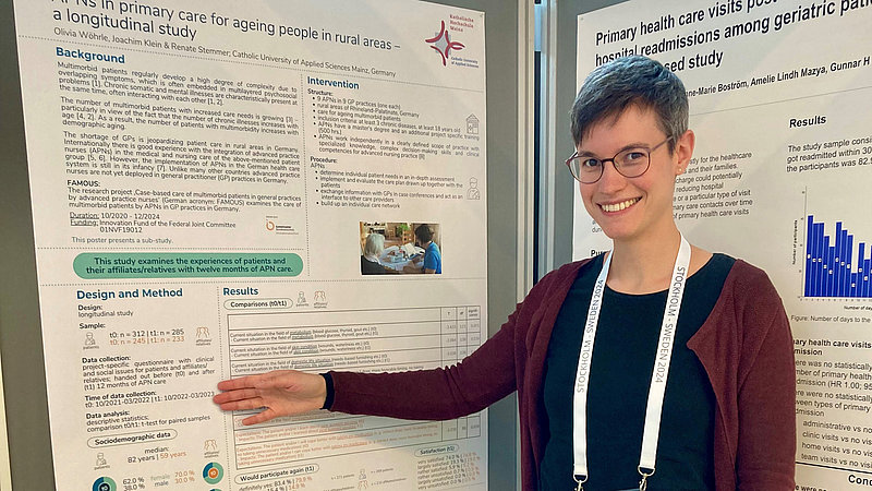 Eine junge Frau deutet auf ein Poster, das an einer Pinnwand hängt und lächelt in die Kamera. 