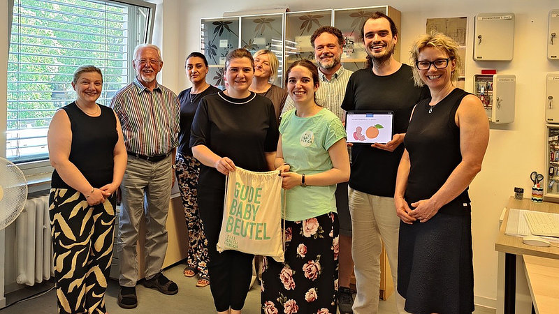 Gruppenbild der Projektbeteiligten, stehend in einem Büro. Ein Frau in der Mitte der Gruppe hält einen Stoffbeutel in der Hand auf dem in hellgrünen Großbuchstaben "Gude Baby Beutel" steht. 
