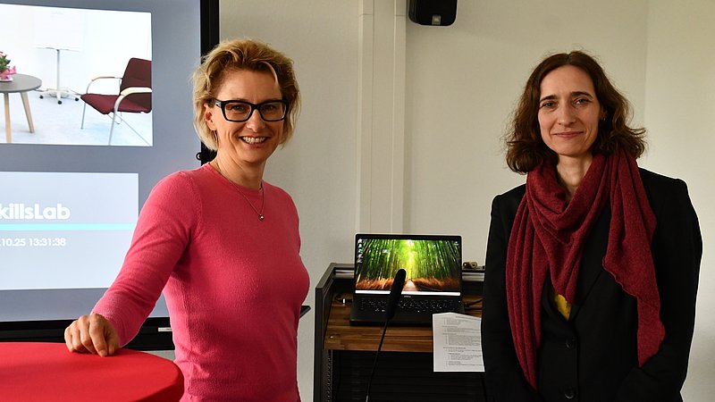 Foto von Professorin Dr. Sandra Bensch und Professorin Dr. Judith Lehnart, die nebeneinander stehend in die Kamera lachen. Im Hintergrund ist die Videotechnik und ein großer Monitor zu stehen 