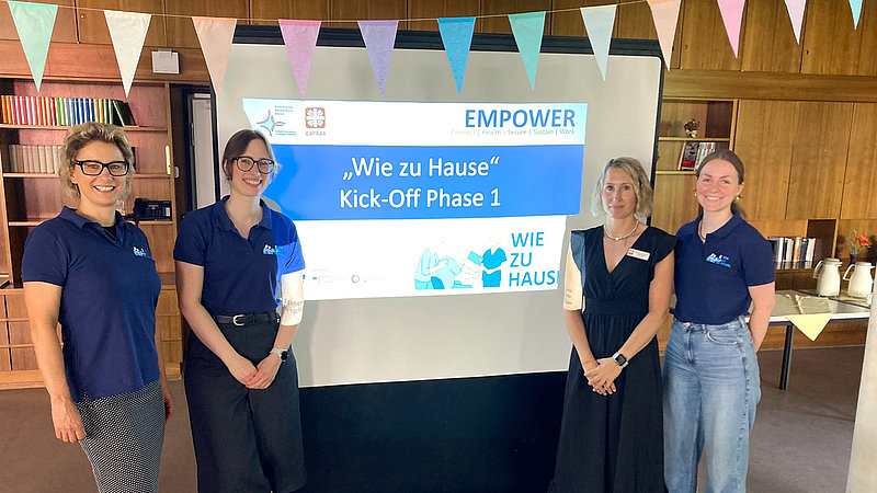 Links und rechts einer Beamer-Leinwand auf der der Projektname "Wie zu Hause " zu lesen ist, stehen jeweils zwei Frauen und lachen in die Kamera.