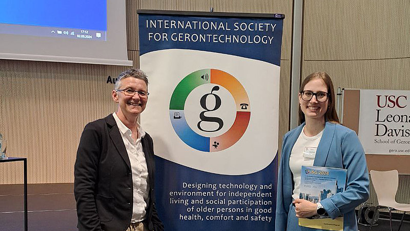 Sabine Corsten und Almut Plath stehen links und rechts von einem Roll-Up mit dem Logo und Schriftzug der International Society for Gerontechnology. 