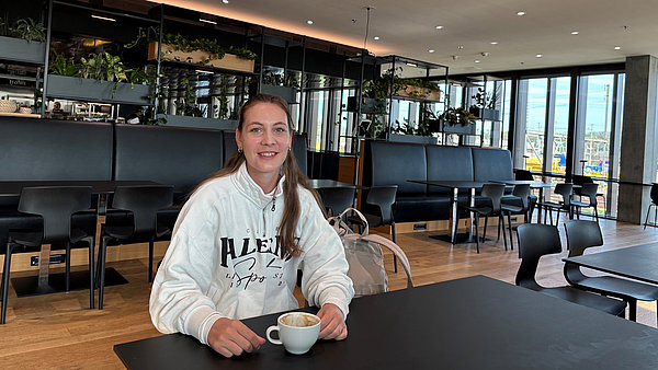 Angela sitzt mit einer Tasse Kaffee vor sich an einem Tisch in einer Cafeteria.