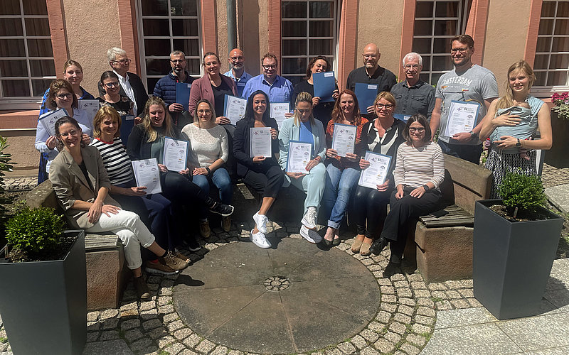 Gruppenbild der Absolventinnen und Absolventen mit ihren Zertifikaten in der Hand. Ein Teil der Gruppe sitzt auf einer labrunden Bank, der andere Teil steht dahinter. 