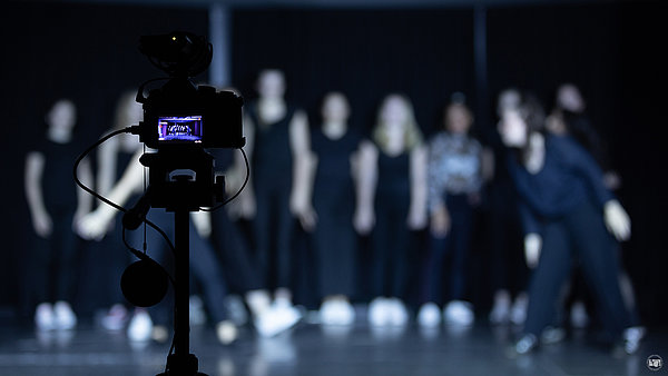 Im Vordergrund steht eine digitale Spiegelreflexkamera. Im Display sieht man eine Gruppe von Personen bei einer Bühnenvorstellung. Die Personen sind im Hintergrund des Bildes unscharf zu erkennen. 
